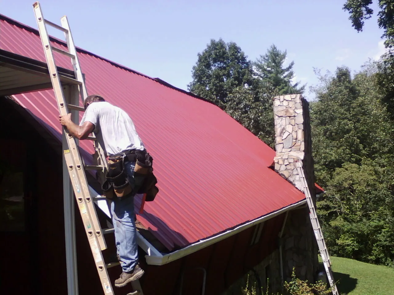 Roofer on a ladder beside a red metal roof and stone chimney — Soffit & Fascia Repair by Roofing Brothers in Cloverdale